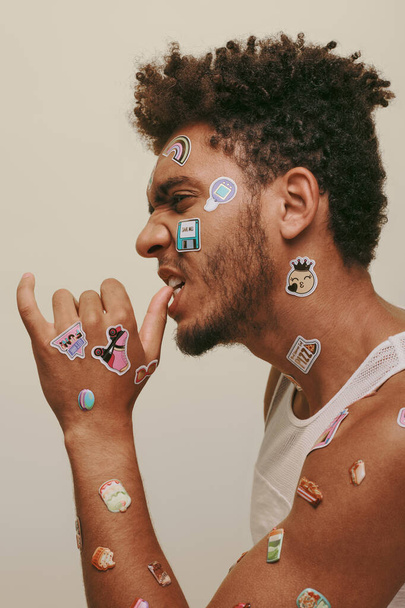 zijaanzicht van Afro-Amerikaanse man met stickers op gezicht kijken naar camera op grijze achtergrond - Foto, afbeelding