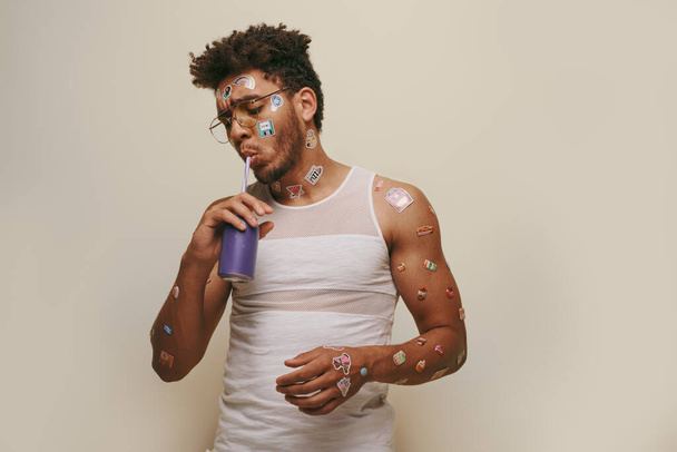 Genç ve kıvırcık Afrikalı Amerikalı adam yüzünde ve vücudunda çıkartmalar olan gri arka planda soda içen. - Fotoğraf, Görsel