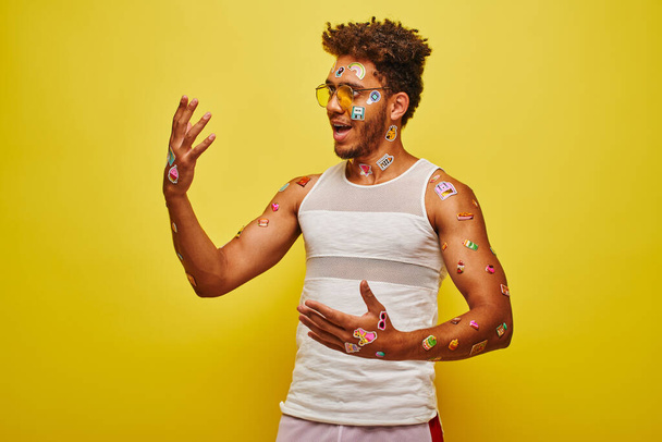 asombrado hombre afroamericano mirando pegatinas en sus manos y cuerpo sobre fondo amarillo - Foto, imagen