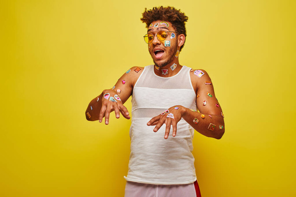 speelse Afro-Amerikaanse man kijkt naar stickers op zijn handen en lichaam op gele achtergrond - Foto, afbeelding
