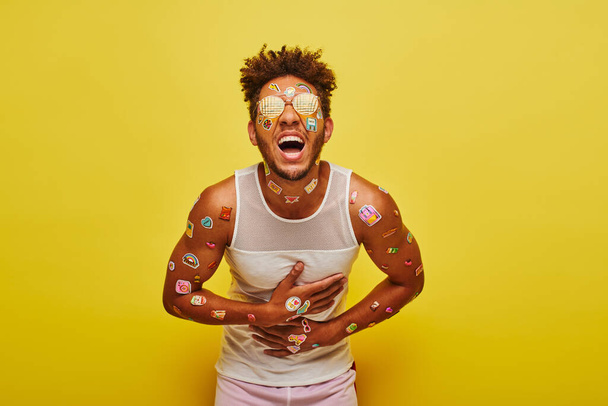 Heyecanlı Afrikalı Amerikalı adam yüzünde ve vücudunda çıkartmalarla sarı arka planda gülüyor. - Fotoğraf, Görsel