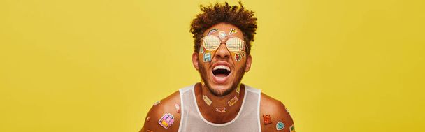 vrolijke Afro-Amerikaanse man met stickers op zijn gezicht en lichaam lachen op gele achtergrond, banner - Foto, afbeelding