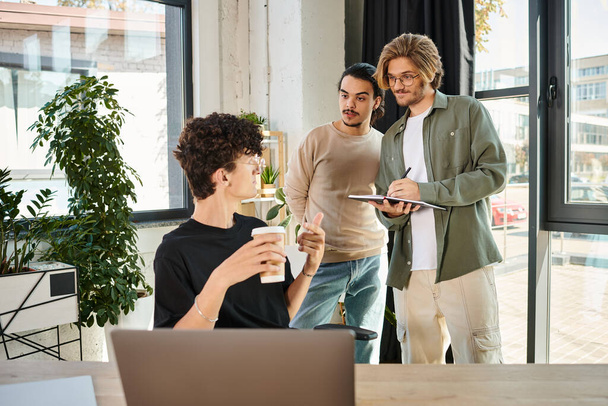Elinde kahve olan genç adam dizüstü bilgisayarı işaret edip proje tartışırken iş arkadaşlarına bakıyor. - Fotoğraf, Görsel