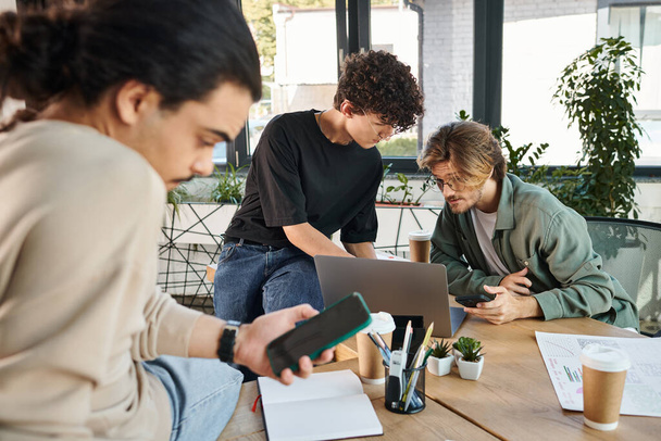 Jovens profissionais brainstorming sobre um laptop no escritório moderno perto de colega de trabalho desfocado com telefone - Foto, Imagem