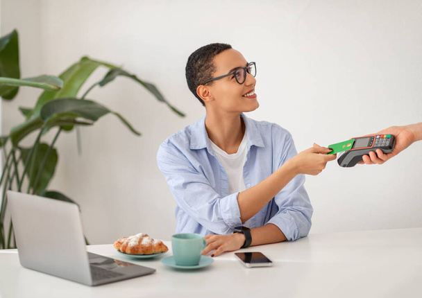Lächelnde junge professionelle Frau, die kontaktloses Bezahlen mit Bankkarte macht, mit modernen Zahlungsterminals in Büroräumen, Cafés. Finanztransaktionen und Technologie, mühelos - Foto, Bild