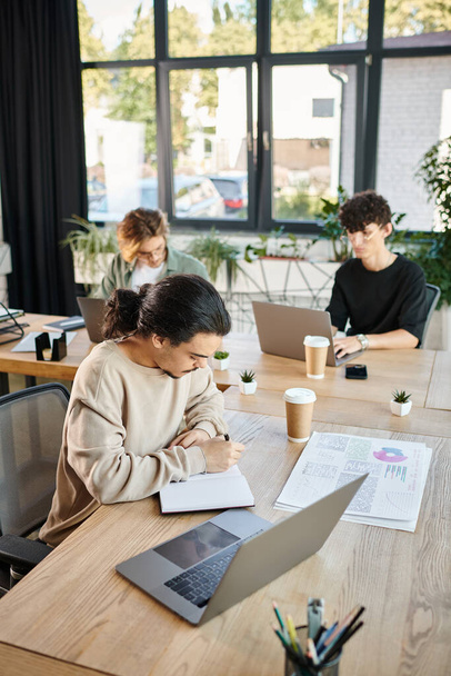 Jeune entrepreneur ciblé analysant les données sur ordinateur portable avec ses collègues dans un espace de travail moderne - Photo, image