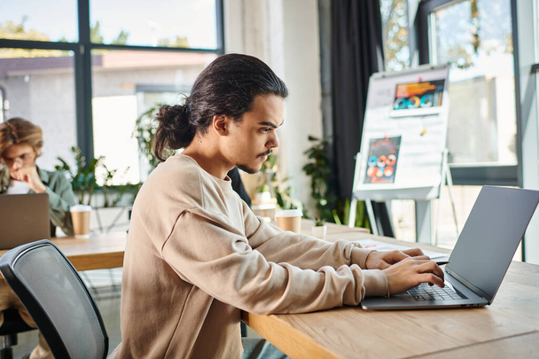 Орієнтований молодий чоловік друкує на ноутбуці з колегами на розмитому фоні в сучасному робочому просторі - Фото, зображення