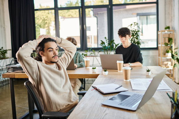 jeune entrepreneur fatigué analyser les données sur ordinateur portable avec des collègues flous dans un espace de travail moderne - Photo, image