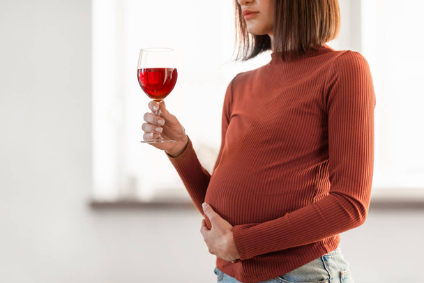 Алкоголь во время беременности. Неузнаваемая молодая беременная женщина, держащая бокал красного вина в помещении дома. Обрезанный снимок беременной женщины, страдающей алкоголизмом - Фото, изображение