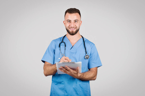 Εύθυμο αρσενικό νοσοκόμα σε μπλε τρίβει κρατώντας σημειώσεις σε ένα πρόχειρο, επιδεικνύοντας προσεκτική τεκμηρίωση της υγειονομικής περίθαλψης και τη διαχείριση της φροντίδας των ασθενών - Φωτογραφία, εικόνα