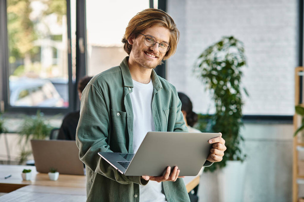 veselý mladý podnikatel v brýlích pomocí notebooku a stojící v moderním coworking prostoru - Fotografie, Obrázek