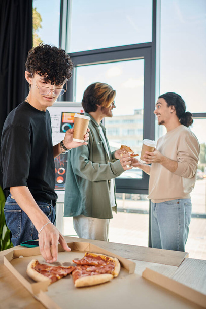 membro da equipe encaracolado com café levando fatia de pizza em ambiente de escritório amigável, pausa para almoço - Foto, Imagem