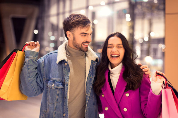 Veselý evropský pár si užívá zimního nakupování, nošení tašek s dárky na slavnostní večer, úsměv v blízkosti městského obchoďáku zdobený světly, objímání a smích při procházce ven - Fotografie, Obrázek
