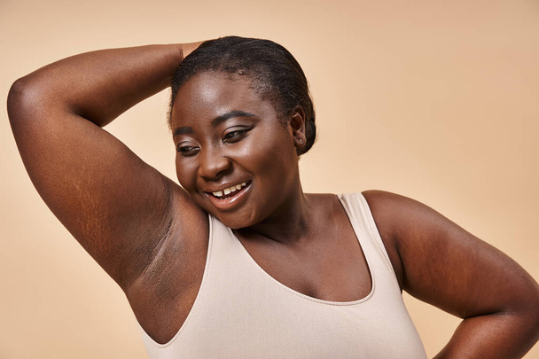 Sebevědomý plus velikost africký americký žena usmívá s rukou za hlavou na béžové pozadí - Fotografie, Obrázek