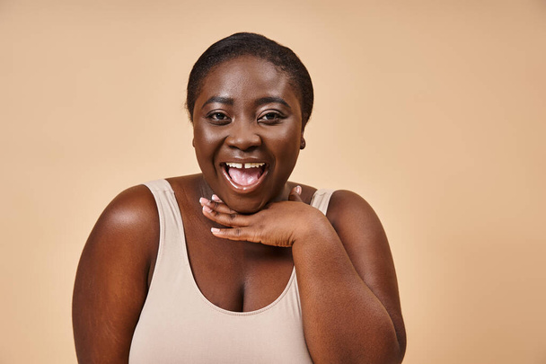 ενθουσιασμένοι συν μέγεθος αφροαμερικανή γυναίκα χαμογελώντας με το χέρι κοντά στο πρόσωπό της σε μπεζ φόντο - Φωτογραφία, εικόνα