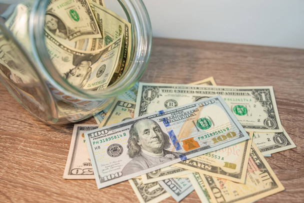 Banknot amerykańskiego dolara leżący na stole, zamknij. Szklana skrzynka pełna pieniędzy w pobliżu, oszczędzając fundusze i koncepcje bogactwa - Zdjęcie, obraz