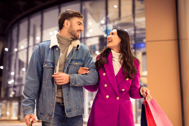 Jeune couple européen aime faire du shopping d'hiver festif, riant ensemble tout en marchant en se tenant la main avec des sacs de cadeaux de vacances, à l'extérieur du centre commercial urbain la nuit d'hiver - Photo, image