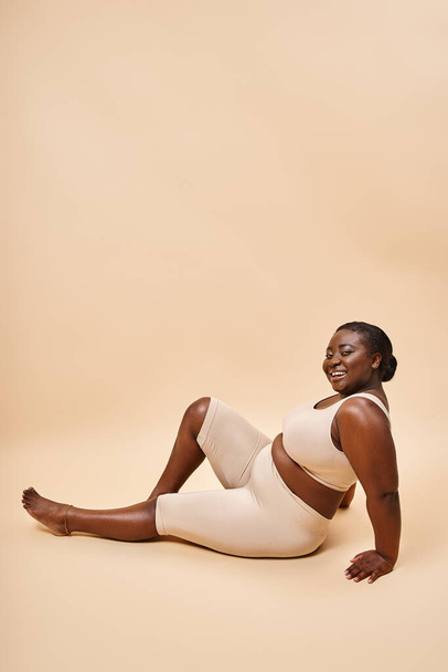 szczęśliwy plus rozmiar afrykański młody kobieta w beżowej bieliźnie pozowanie na pasujące tło - Zdjęcie, obraz