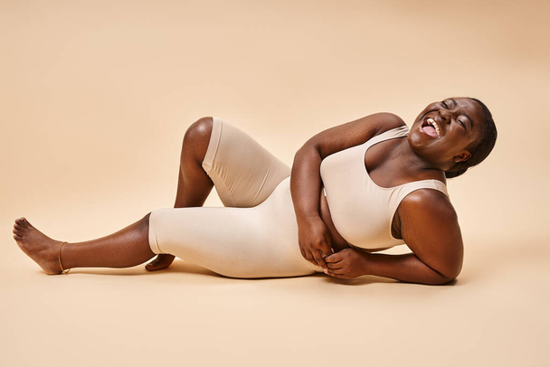 plus size mulher americana africana em lingerie bege rindo e reclinado contra o pano de fundo correspondente - Foto, Imagem