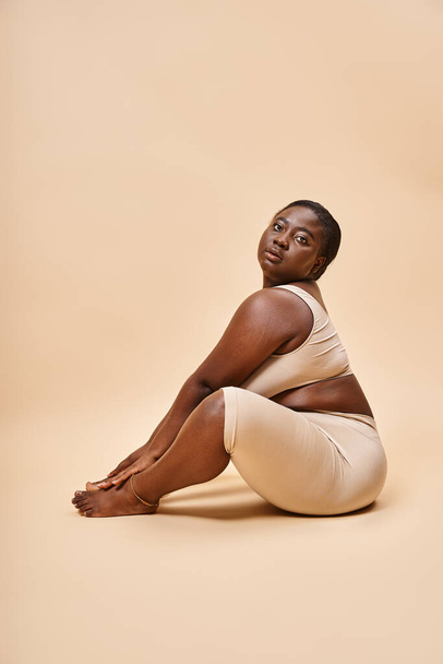 cuerpo positivo, mujer joven afroamericana en ropa interior beige posando contra el telón de fondo a juego - Foto, Imagen