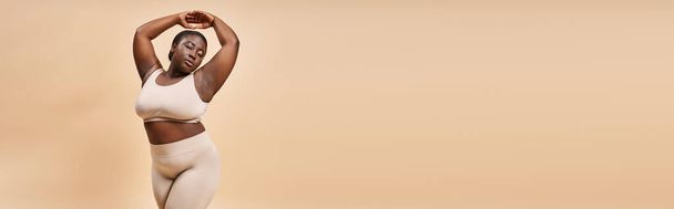 plus size donna afroamericana in biancheria intima posa con le mani sollevate su sfondo beige, banner - Foto, immagini