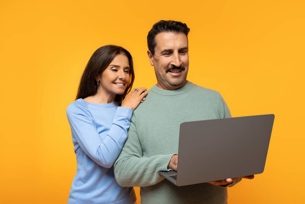 Lächelnder europäischer Mann in Pullover und Frau schauen gemeinsam auf den Laptop-Bildschirm, Dame stützt Hand auf seine Schulter, beide zeigen Ausdruck von Interesse und Vergnügen auf orangefarbenem Hintergrund - Foto, Bild