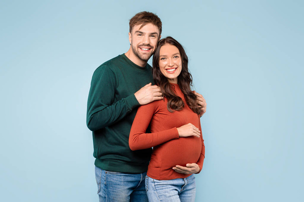 放射性期待カップルは,一瞬を共有し,男は愛情を持って彼の妊娠中のパートナーを抱擁し,どちらも幸せに青い背景に微笑みます - 写真・画像