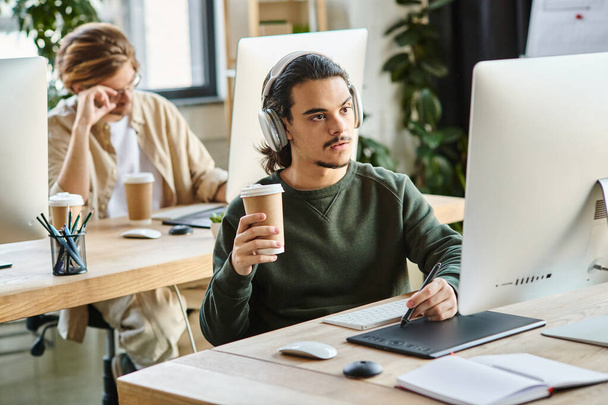 zamyślony mężczyzna profesjonalista w słuchawkach trzymających kawę iść i za pomocą tabletu graficznego w biurze - Zdjęcie, obraz