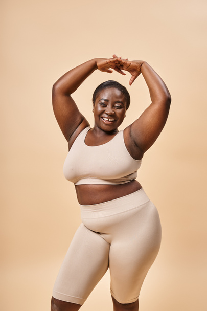 happy plus size woman in beige underwear posing with raised hands, body positive and self esteem - Foto, imagen