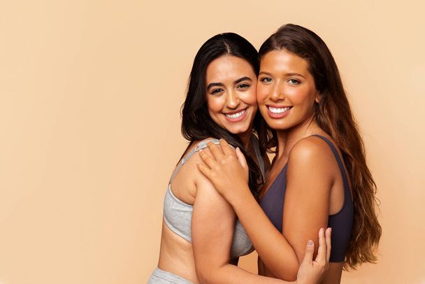 Two diverse positive young women in underwear, hugs, enjoy body care, isolated on beige studio background. Wellness, friendship, spa procedures, fit ad and offer, lifestyle - Foto, imagen