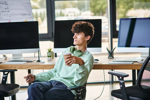 Genç kıvırcık saçlı adam ofis koltuğunda oturuyor ve elinde kahve tutuyor. - Fotoğraf, Görsel