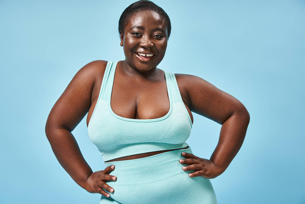 happy plus size afrikanisch-amerikanische Frau in blauer Sportbekleidung mit Händen auf Hüften vor passendem Hintergrund - Foto, Bild
