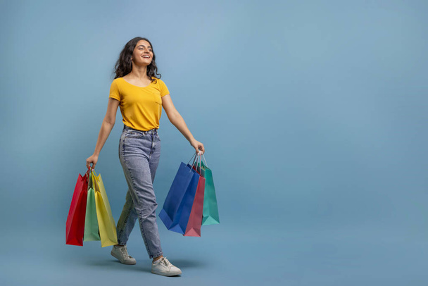 Happy joyful young eastern woman student walking with colorful shopping bags in her hands towards copy space for advertisement, isolated on blue studio background. Shopomania, sale season - Photo, Image
