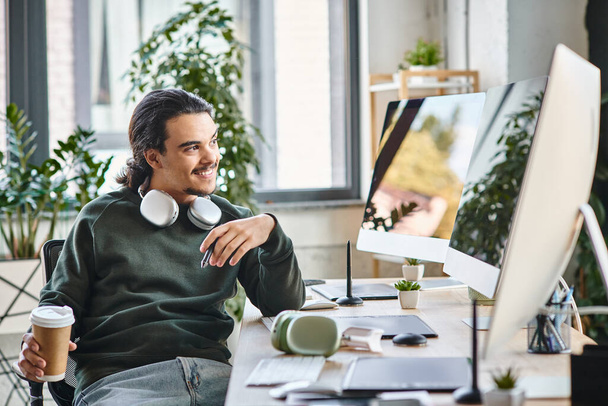 Młody człowiek z długopisem i kawą uśmiechający się i patrzący na monitor w poprodukcyjnej przestrzeni roboczej - Zdjęcie, obraz