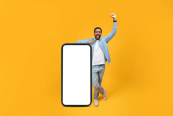 Uomo estatico in abbigliamento casual tifo e indicando un mockup smartphone oversize vuoto accanto a lui, su uno sfondo giallo monocromatico. Raccomandazione app - Foto, immagini