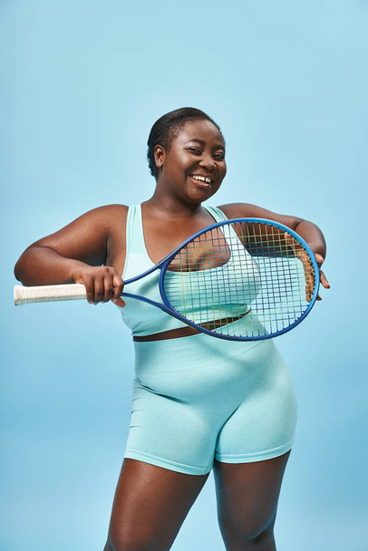 šťastný plus velikost tmavé kůže žena drží tenisovou raketu na modrém pozadí, tělo pozitivní a sport - Fotografie, Obrázek