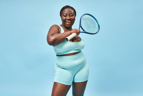 Neşeli ve esmer tenli bir kadın mavi arka planda tenis raketi tutuyor, vücut pozitifliği var. - Fotoğraf, Görsel