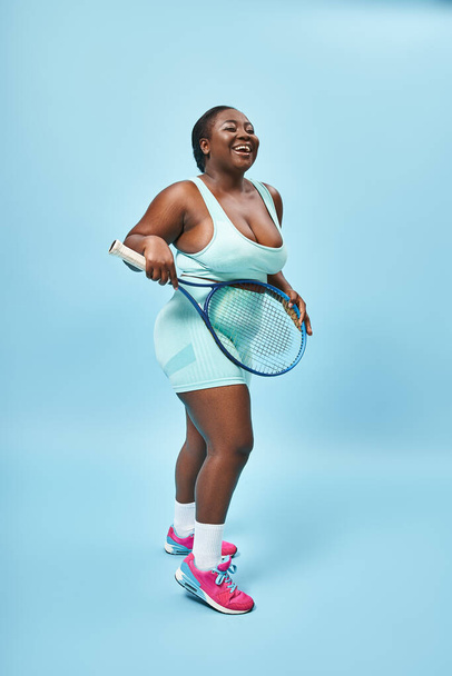 feliz más tamaño de piel oscura mujer sosteniendo raqueta de tenis sobre fondo azul, cuerpo positivo y deporte - Foto, imagen