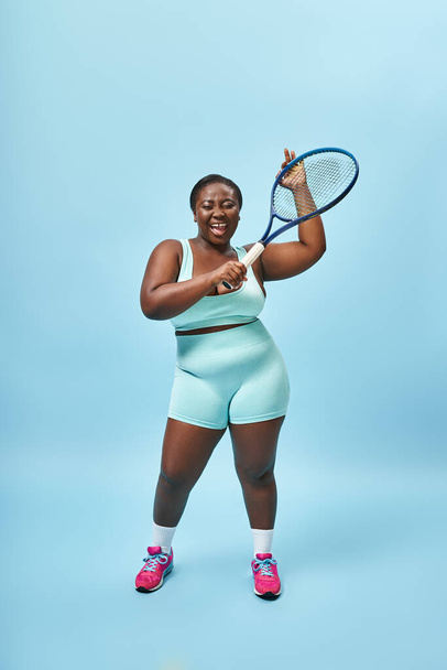 excited plus size dark skinned sportswoman holding tennis racket on blue backdrop, body positive - Photo, Image