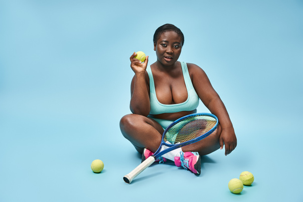 joven deportista de piel oscura más tamaño sosteniendo raqueta de tenis y sentado alrededor de las bolas en azul - Foto, imagen