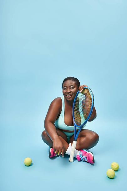Mutlu ve esmer tenli kadın tenis raketi tutuyor ve mavi topların etrafında oturuyor. - Fotoğraf, Görsel