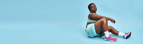 Χαλαρωμένη συν μέγεθος Αφροαμερικανή γυναίκα κάθεται σε μπλε αθλητικά ρούχα σε φόντο που ταιριάζουν, πανό - Φωτογραφία, εικόνα