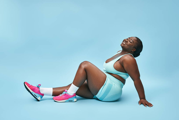 ontspannen plus size Afrikaanse Amerikaanse vrouw zitten in blauwe sportkleding op bijpassende achtergrond, glimlach - Foto, afbeelding