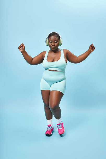 Radostné plus velikost africké americké ženy tančí v aktivním oblečení se sluchátky na, modré pozadí - Fotografie, Obrázek