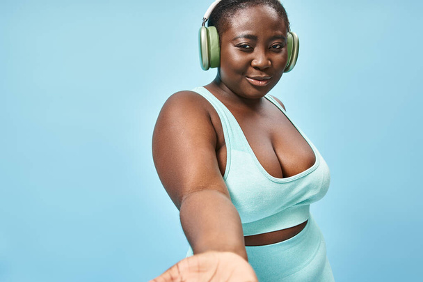 щасливий плюс розмір афроамериканської жінки в активному одязі, слухаючи музику в навушниках, синій фон - Фото, зображення