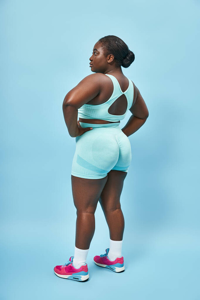 plus velikost africká americká žena ve stylovém aktivním oblečení při pohledu zpět a pózování s rukama v bok - Fotografie, Obrázek