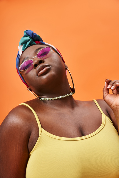 portrait of stylish plus size african american woman in headscarf and trendy sunglasses on orange - Photo, Image
