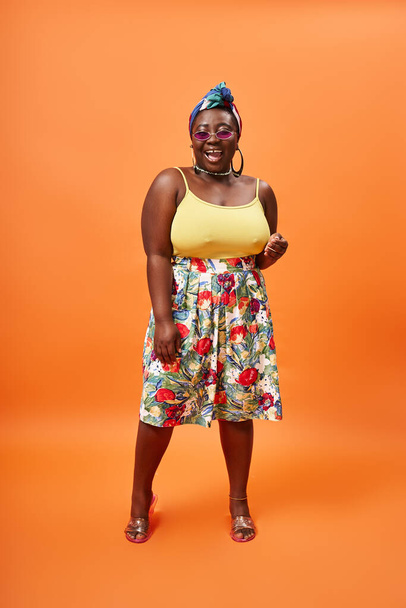 animado afro-americano plus size mulher com saia floral e óculos de sol posando em pano de fundo laranja - Foto, Imagem