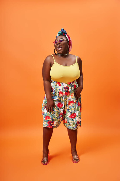 jolly african american plus size woman with floral skirt and sunglasses posing on orange backdrop - Photo, Image