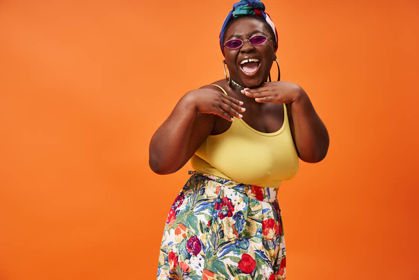 vzrušený africký Američan plus velikost žena s šátkem a slunečními brýlemi pózovat na oranžovém pozadí - Fotografie, Obrázek
